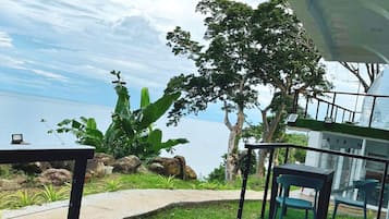 Una terraza con vista al mar, que incluye una mesa y sillas, rodeada de follaje y rocas.