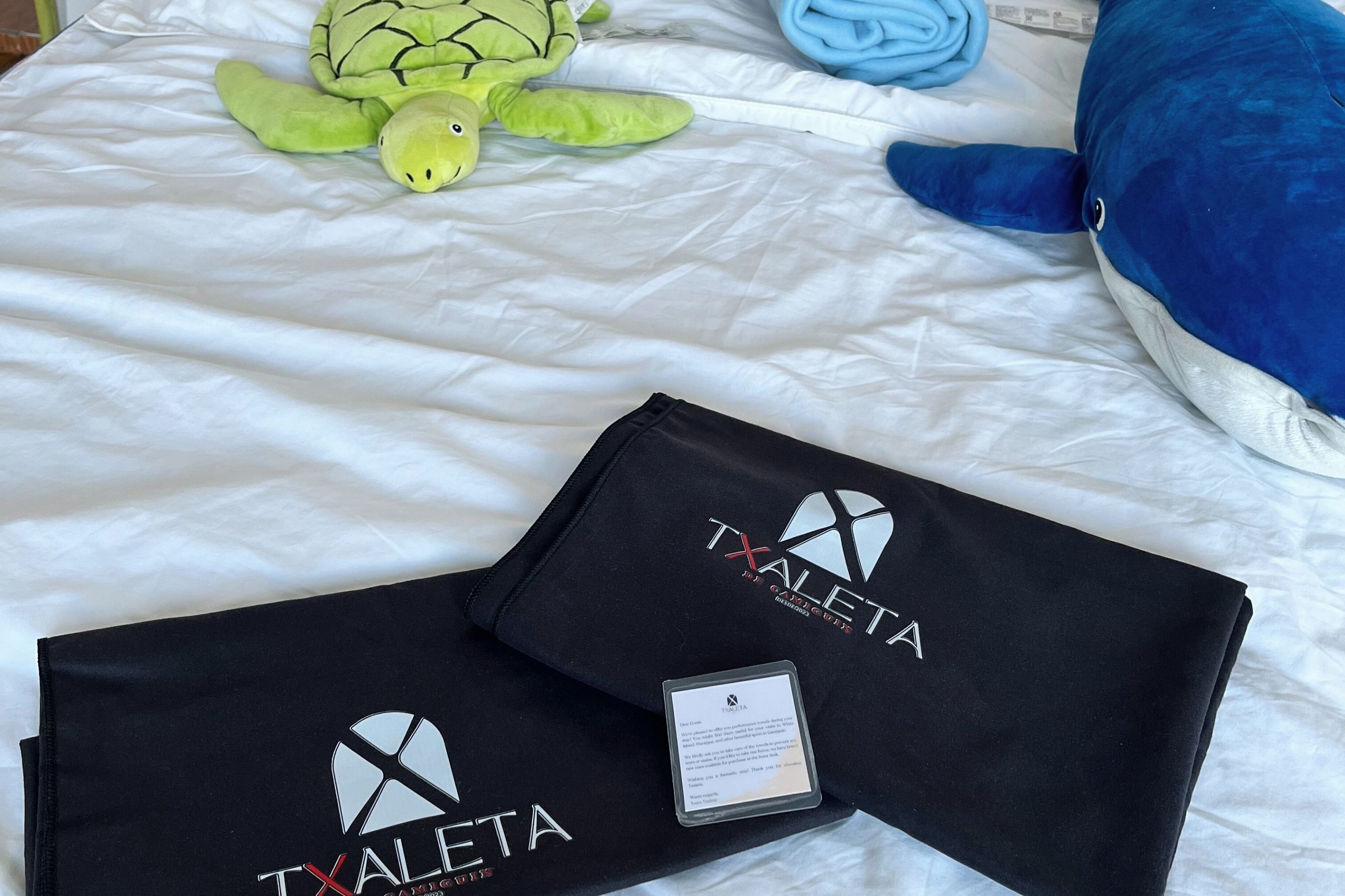 Una cama con sábana blanca, un peluche de tortuga verde y dos toallas negras con el logotipo 'TALETA'.