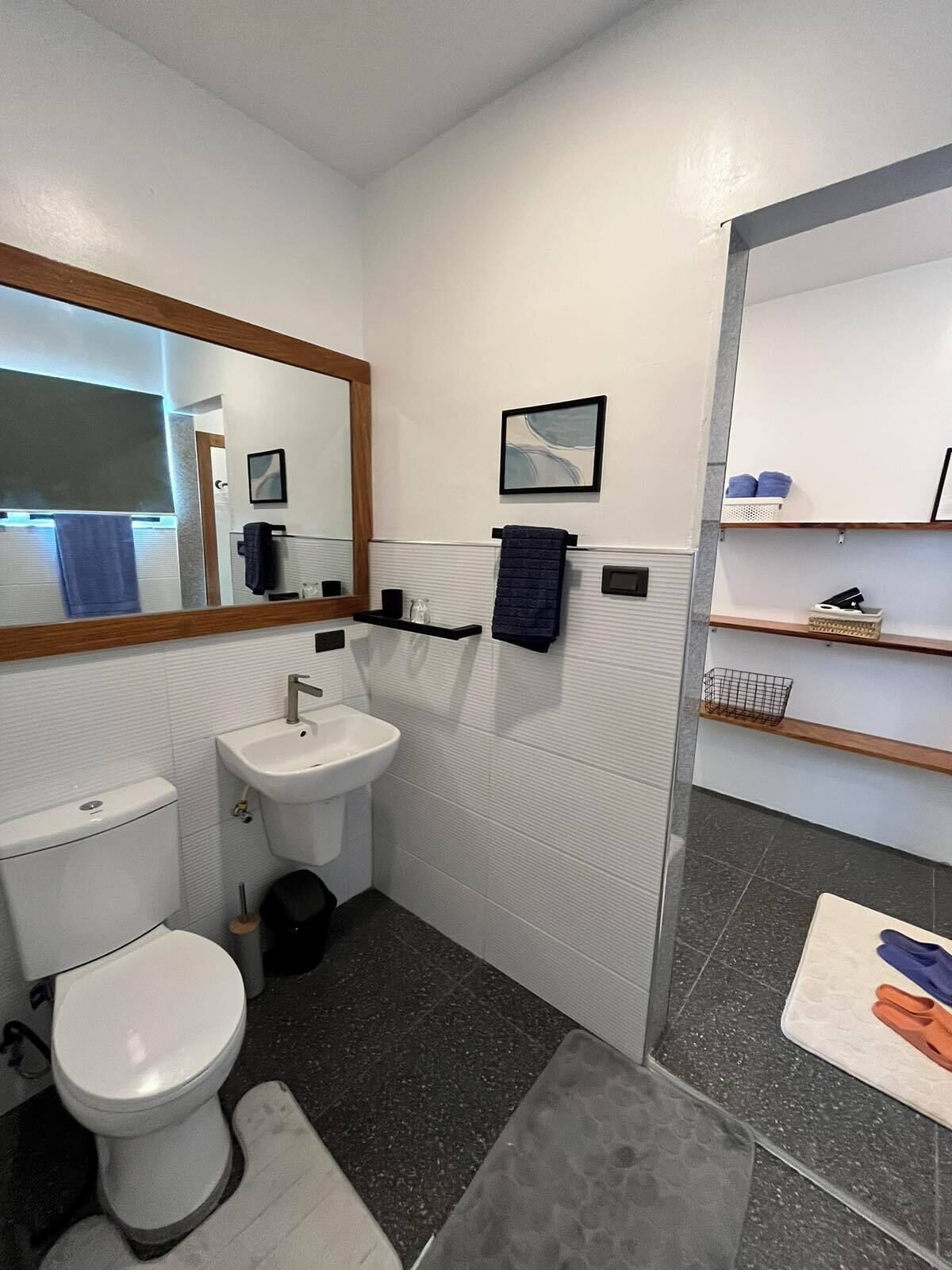 A modern bathroom with a toilet, sink, and mirror. There are shelves with towels and toiletries.