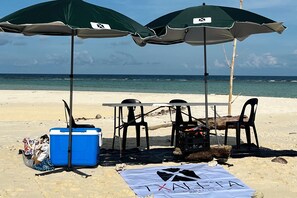 Strandaufstellung mit zwei Sonnenschirmen, einem Tisch, Stühlen, einer Kühlbox und einem Handtuch mit dem Schriftzug 'TAXA'.
