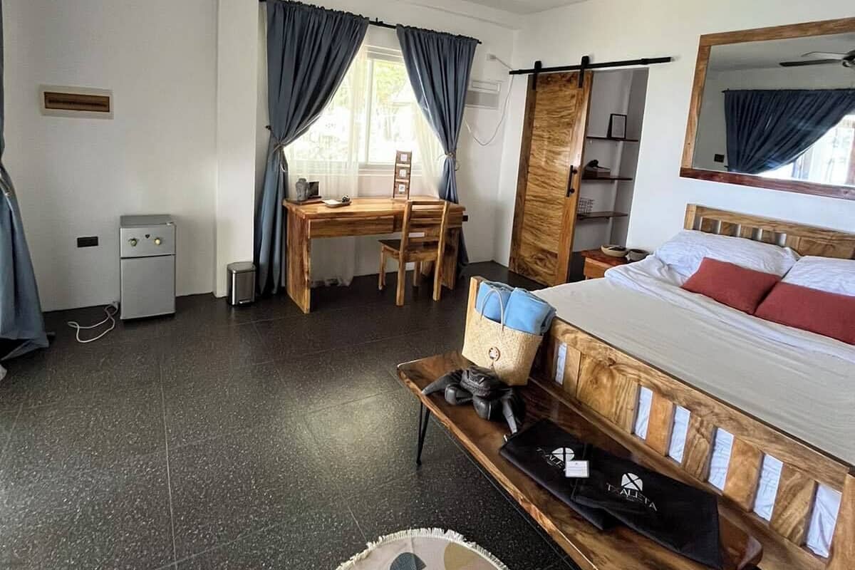 Ein Hotelzimmer mit einem Bett, einem Schreibtisch, einem Stuhl, einem kleinen Tisch, einem Deckenventilator und einem fenster mit Vorhängen.