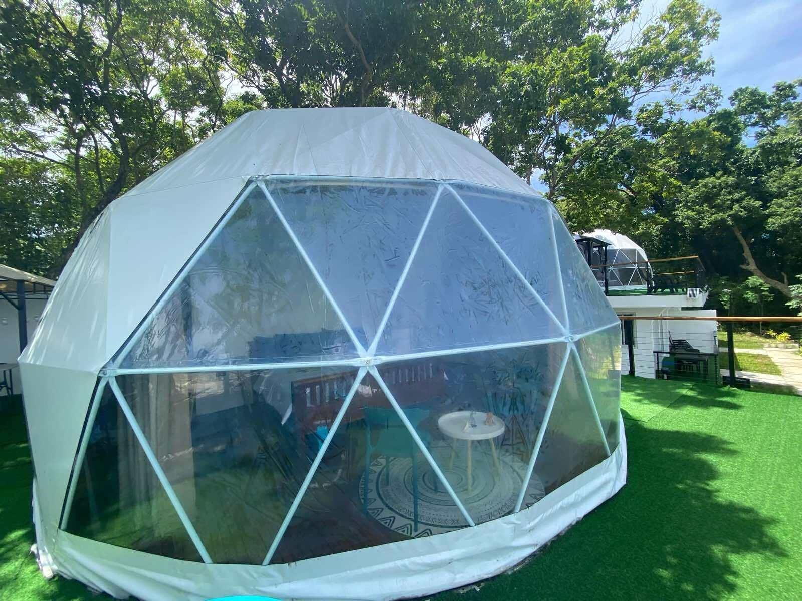 A geodesic dome structure with a transparent roof, surrounded by green grass and trees.