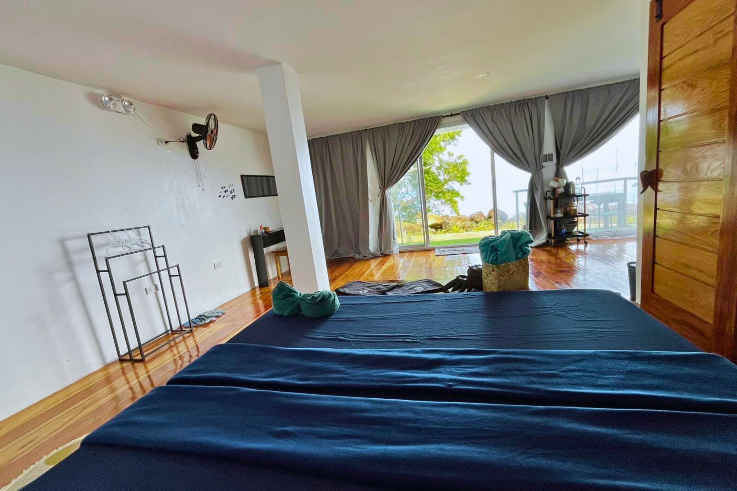A spacious bedroom with a large bed, a wooden floor, a balcony with a view of greenery, and a ceiling fan.