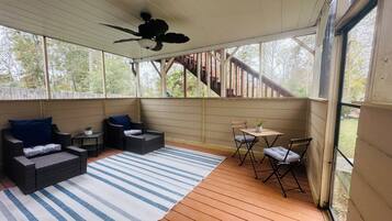 Un espace extérieur abrité, doté d'un ventilateur de plafond, de meubles en osier, d'un tapis à rayures et offrant une vue sur une terrasse en bois et des arbres.