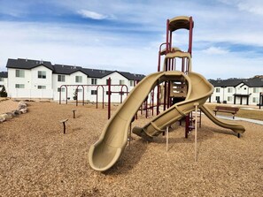 A playground with slides and climbing structures, surrounded by a gravel area and residential buildings in the background.
