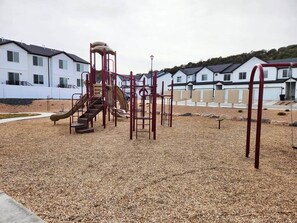A playground with climbing structures and slides, surrounded by a gravel area and residential buildings in the background.