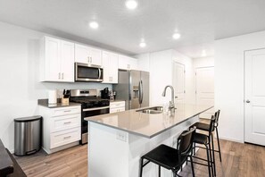 A modern kitchen with white cabinets, stainless steel appliances, a central island with bar stools, and a trash can.