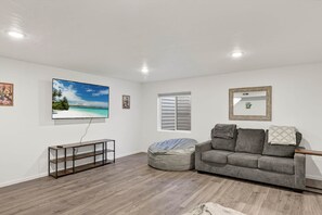 A living room with a flat-screen TV, a gray sofa, a bean bag, a wooden coffee table, and a window with shutters.