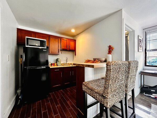 Una cocina compacta con armarios de madera oscura, un refrigerador negro y una silla de fibras naturales.