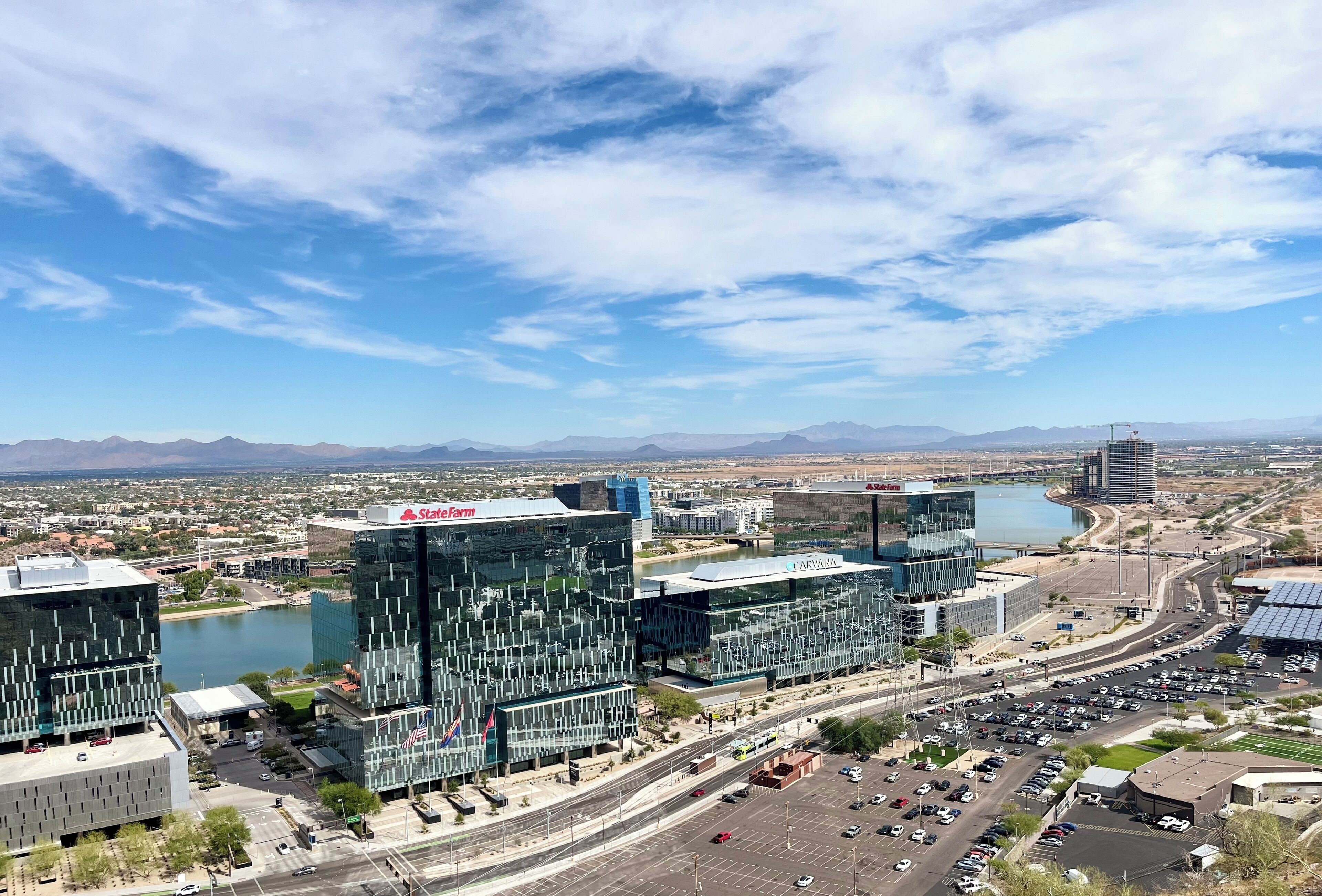A modern cityscape with prominent buildings, including a large glass structure with a red logo, a body of water, and a highway with vehicles.