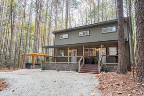 A two-story cabin with a covered porch and a stone foundation, surrounded by a forest. - Pine Cone Cottage - Pine Cone Cottage (Hamptonville)