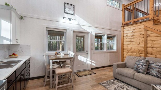 Une cuisine avec des meubles blancs, une table à manger avec des chaises, un canapé et un escalier.