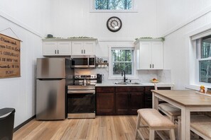 A modern kitchen with stainless steel appliances, white cabinetry, and a wooden dining table. - Pine Cone Cottage - Pine Cone Cottage (Hamptonville)