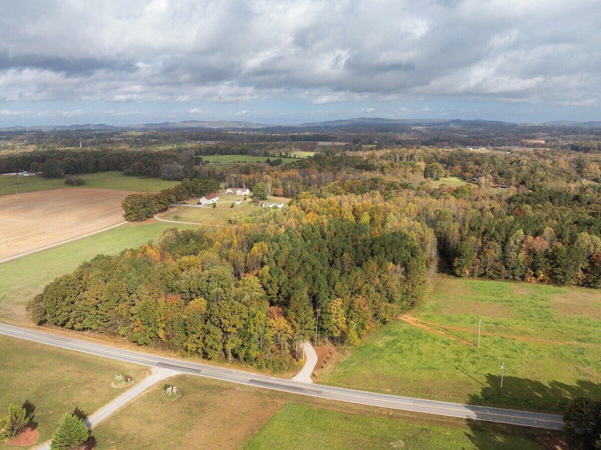 Une vue aérienne d’un paysage rural, avec une route, des champs et un ensemble de bâtiments entourés d’arbres.