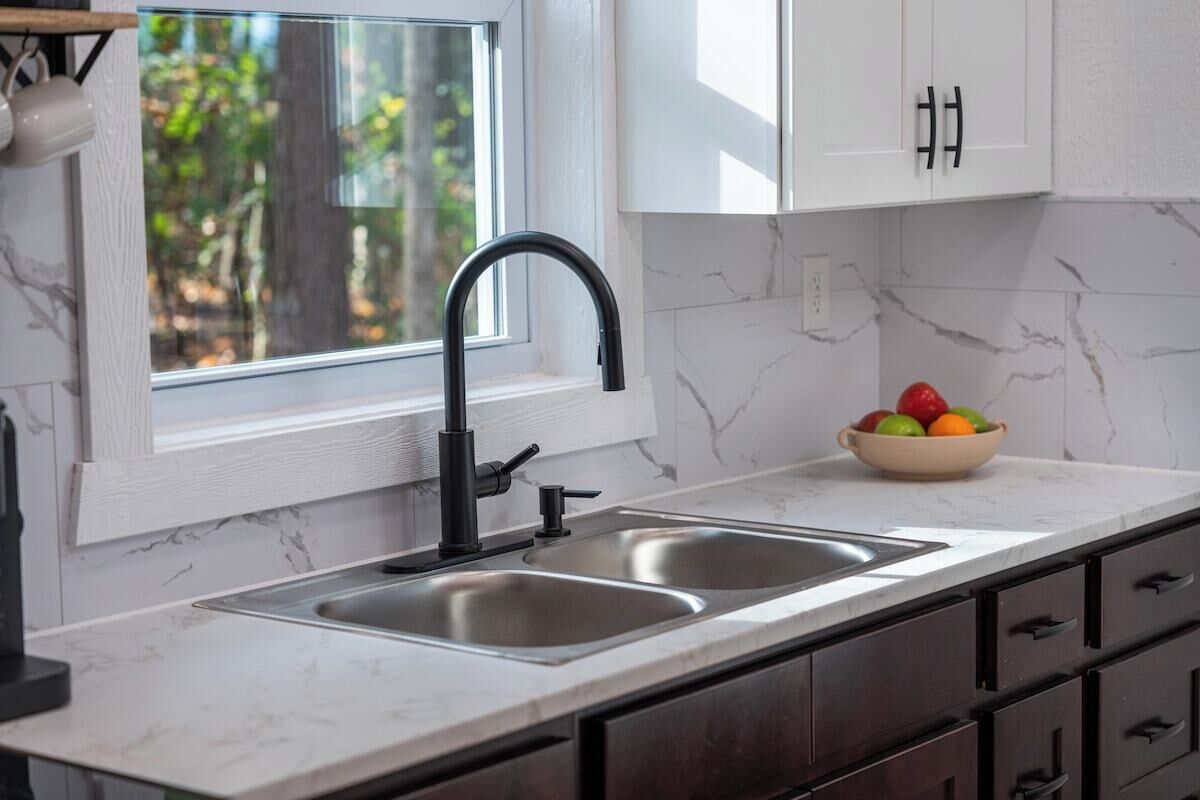 A kitchen with a double sink, white cabinets, and a window with a view of trees.