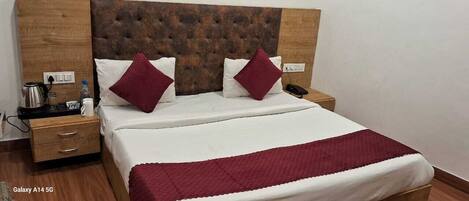 Un lit bien fait, recouvert d’un plaid rouge, une table de chevet en bois avec une bouilloire et un téléphone, et une tête de lit marron à motifs capitonnés.