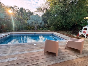 Ein Schwimmbecken mit zwei Liegen auf einer Holzterrasse, umgeben von üppigem Grün.