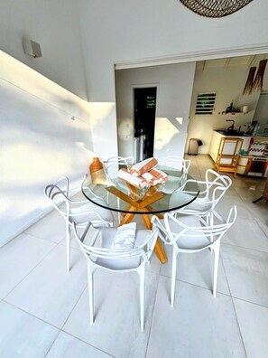Une table ronde en verre, entourée de chaises blanches, un coin cuisine avec une cuisinière et des placards, et une porte donnant sur une autre pièce.