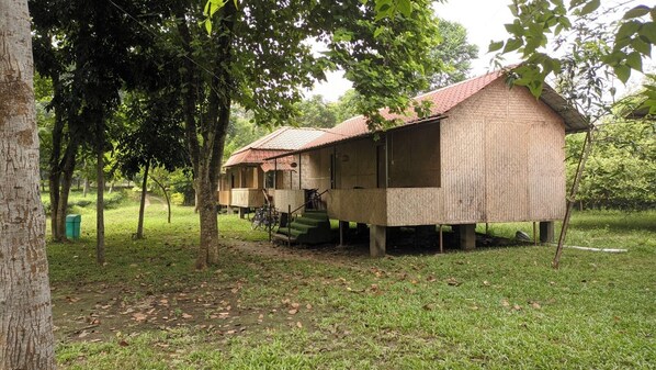 A rustic wooden structure with a tiled roof, elevated on stilts, surrounded by trees and grass.