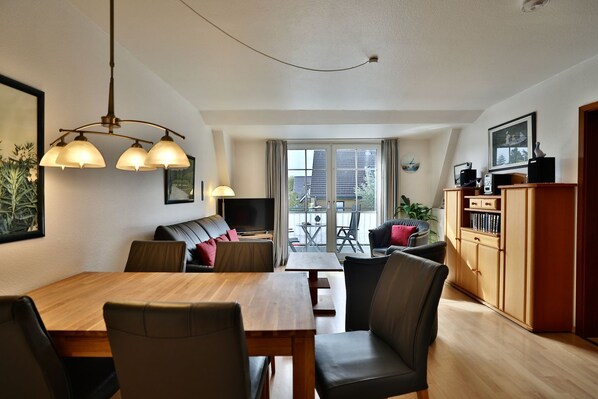A modern living room with a dining table, sofas, and a TV. There is a balcony with a view of buildings.