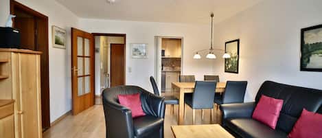A modern living room with a black leather sofa, a wooden coffee table, and a dining area with a wooden table and chairs.