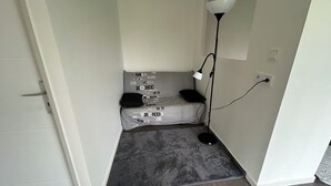 Una stanza piccola e stretta con tappeto grigio, una porta bianca, una lampada da terra nera e un letto con biancheria grigia.