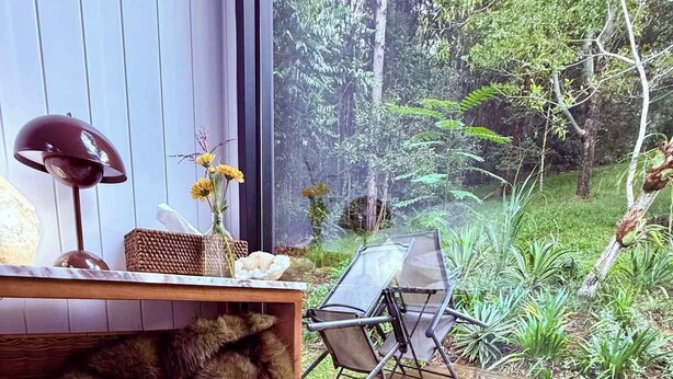 Een kamer met een groot raam, een rieten mand, een fauteuil met een bontdierenbekleding en een kleine tafel met een lamp.