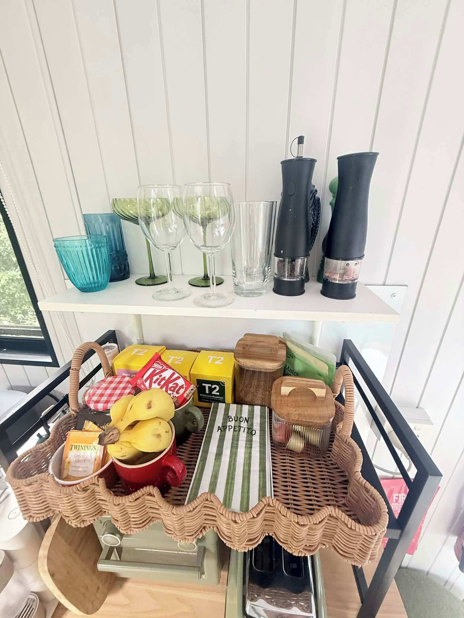 Una cocina con una pizarra, una cesta de mimbre con alimentos y un estante con cristalería y utensilios de cocina.