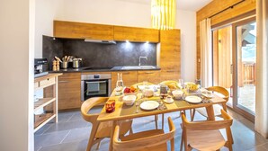 A modern kitchen with wooden cabinets, a dining table set for four, and a large window with curtains. - APARTMENT  CHÂTEL - O Rouge A204 (CHATEL)
