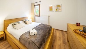 A bedroom with a wooden bed, a nightstand with a lamp, a wooden dresser, and a window with curtains. - APARTMENT  CHÂTEL - O Rouge A204 (CHATEL)