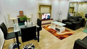 A modern living room with a glass dining table, black and white chairs, a white coffee table, a black sofa, and a TV mounted on the wall.
