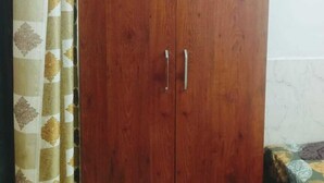 A wooden wardrobe with two doors and handles, positioned next to a bed with a patterned bedspread. - Shree Sitaram Guest House (Faizabad)