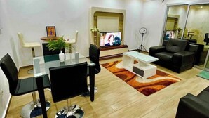 Ein modernes Wohnzimmer mit einem Glastisch, schwarzen und weißen Stühlen, einem weißen Couchtisch, einer schwarzen Couch und einem an der Wand montierten Fernseher.