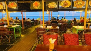 A restaurant with tables and chairs, a wooden floor, and a view of the ocean. - Rainbow Resort Bar and Restaurant (Morjim)