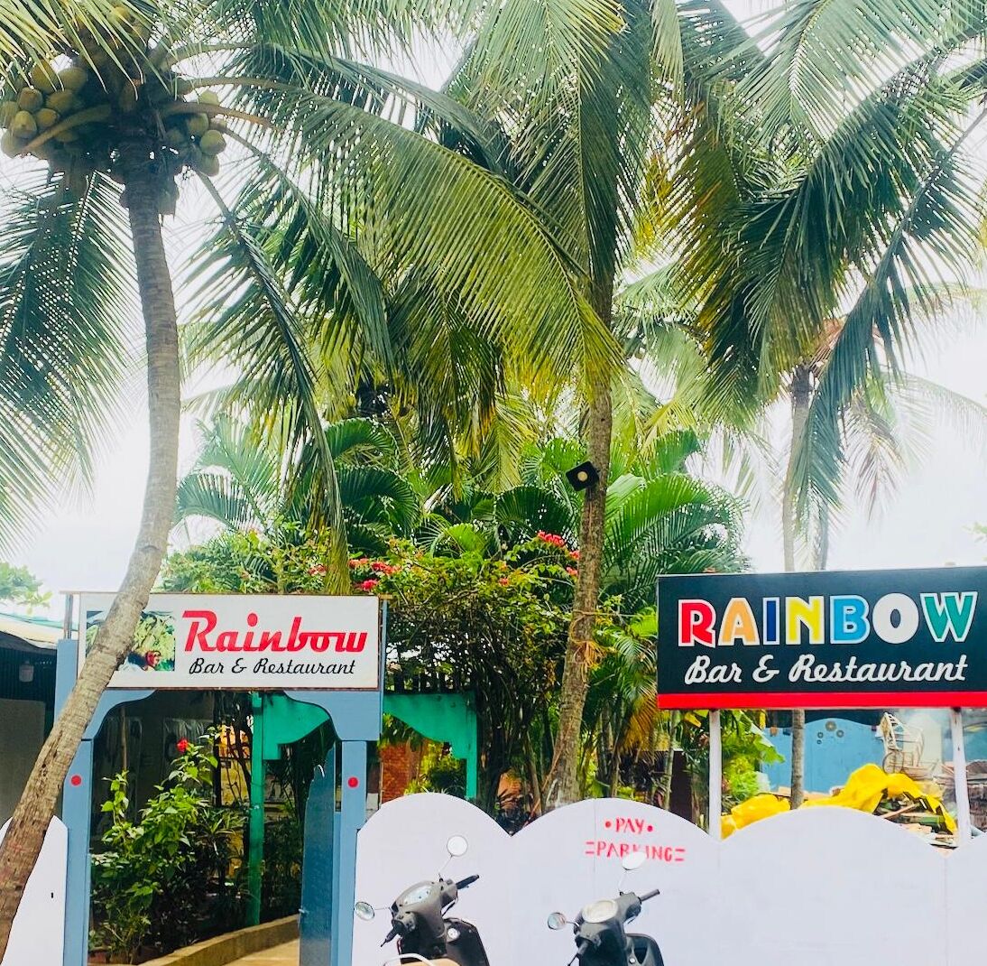 A restaurant named 'Rainbow Bar & Restaurant' with a signboard, surrounded by lush greenery and palm trees.