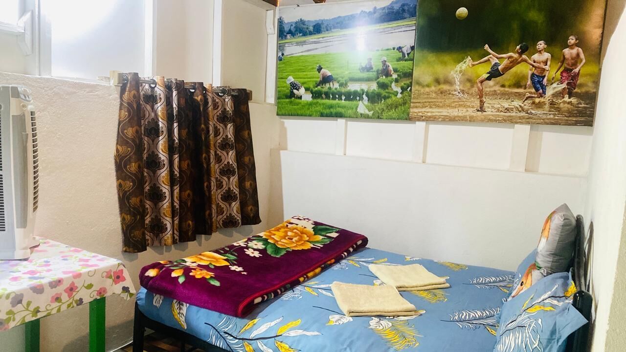 Una camera da letto con un letto, un tavolo con tovaglia a fiori, una finestra con tende e una parete con un quadro incorniciato.