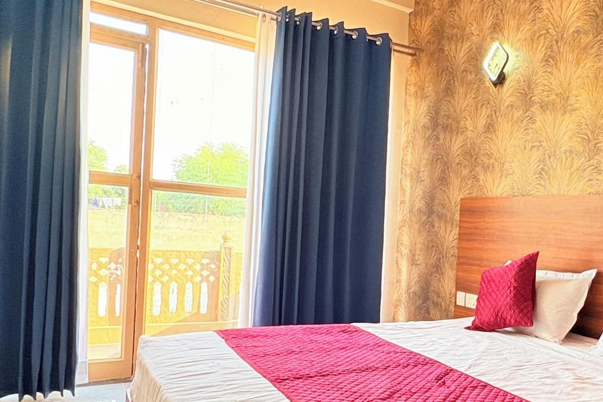 A hotel room with a large bed, a wooden headboard, a red and white quilted bedspread, and a view of a garden through a sliding glass door.