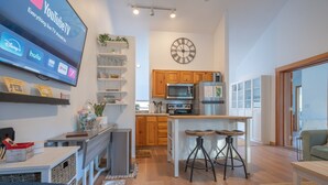A modern kitchen with wooden cabinets, a central island with stools, a flat-screen TV mounted on the wall, and a clock above the cabinets. - Tolovana 3 - 700ft to Beach - Family Fun - Dogs Ok! (Cannon Beach)