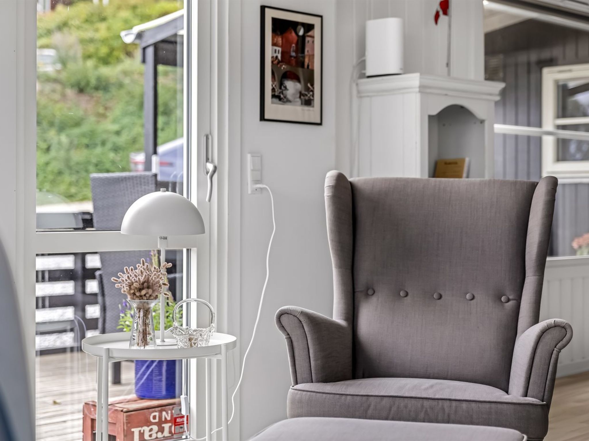 Een grijs bekleden fauteuil in een kamer met een witte muur, een ingelijst schilderij en een kleine ronde tafel met een vaas en een mand.