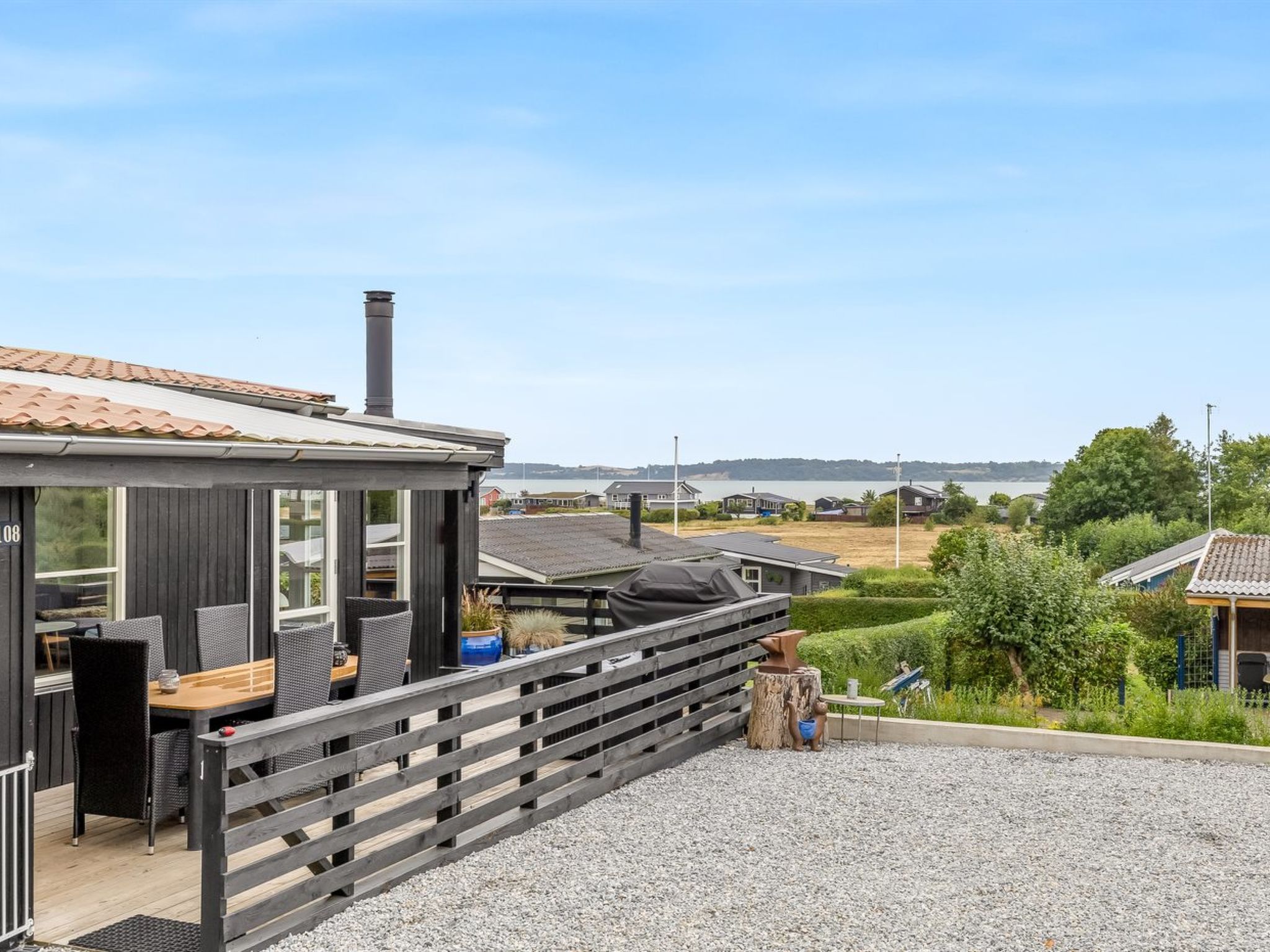 Un área de estar moderna al aire libre con una terraza de madera, una parrilla y vista a un lago y edificios residenciales al fondo.