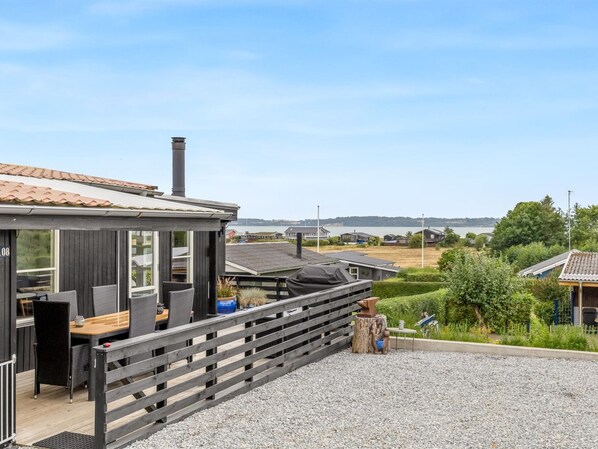 A modern outdoor seating area with a wooden deck, a barbecue grill, and a view of a lake and residential buildings in the background.