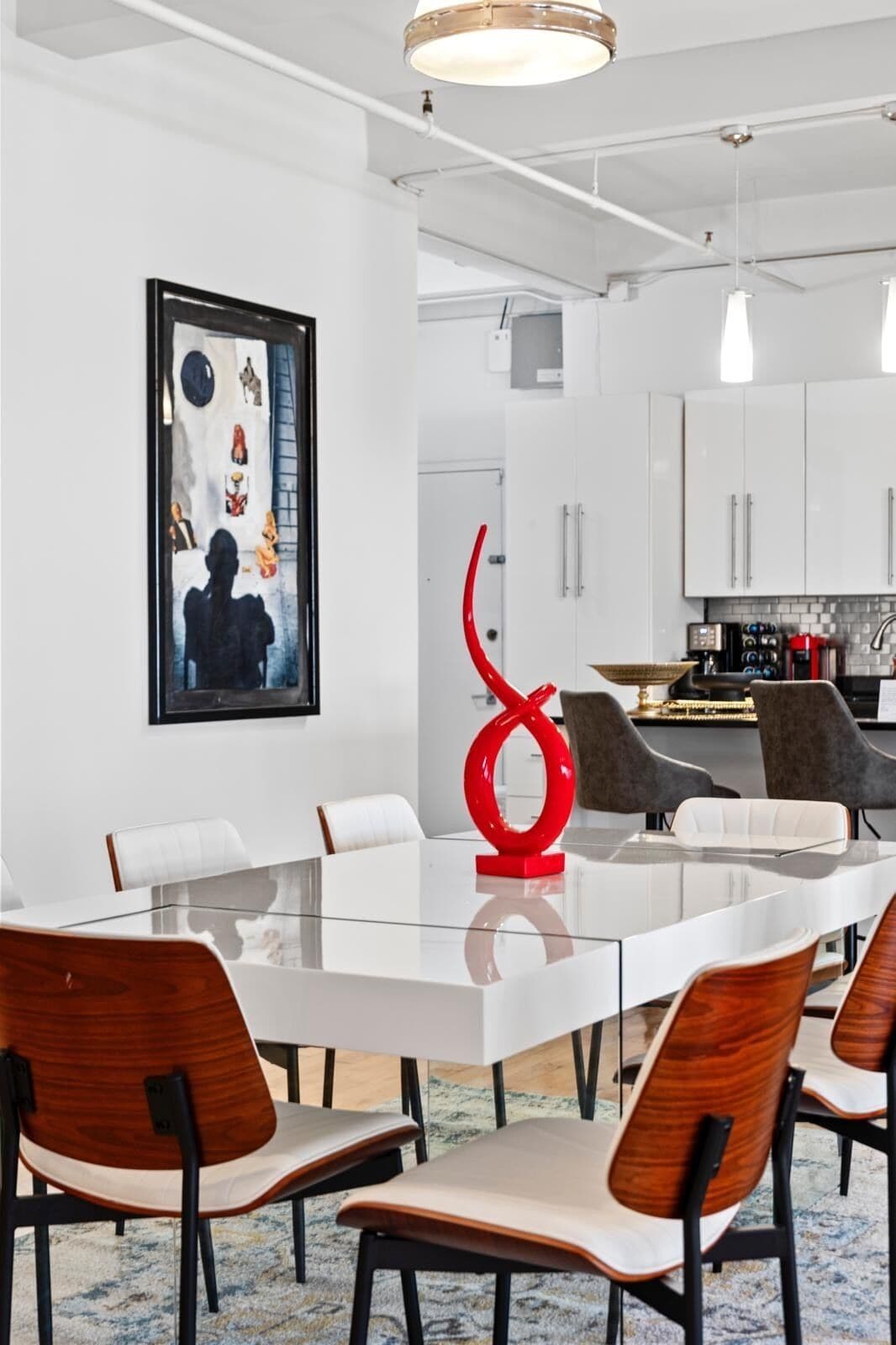 A modern dining area with a glass table, wooden chairs, a red abstract sculpture, and a framed artwork on the wall.