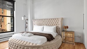 Une chambre à coucher avec une tête de lit capitonnée, un lit rond assorti d’un pouf, une table de chevet agrémentée d’un vase et une fenêtre donnant sur un immeuble.