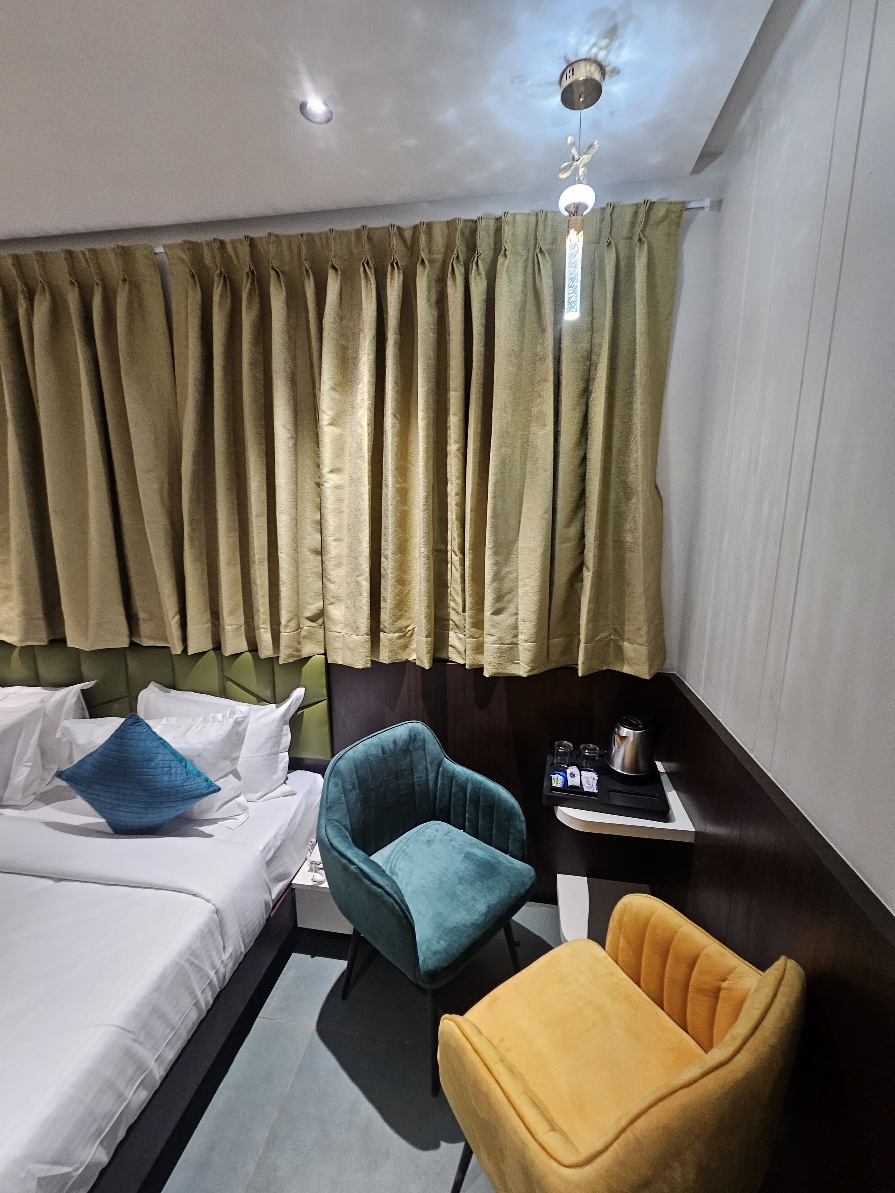 Una habitación de hotel con una cama, dos sillas, una mesita con hervidor y cortinas.
