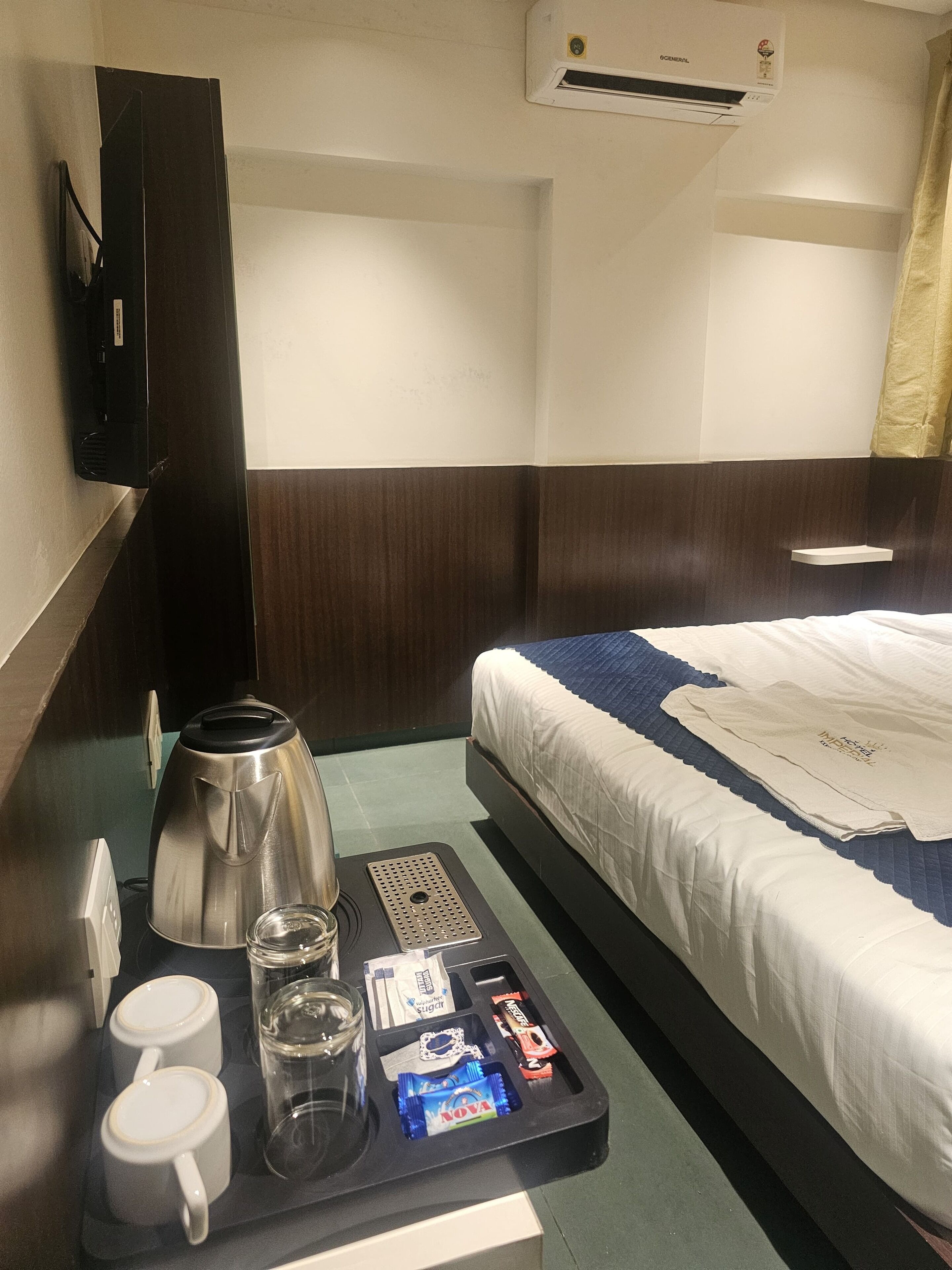 A hotel room with a bed, a bedside table with a coffee maker, cups, glasses, and snacks.