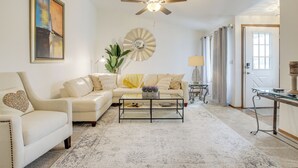 Un salon spacieux doté d’un ventilateur de plafond, d’un grand tableau abstrait, d’un miroir rond et d’un canapé agrémenté d’un coussin en forme de cœur.