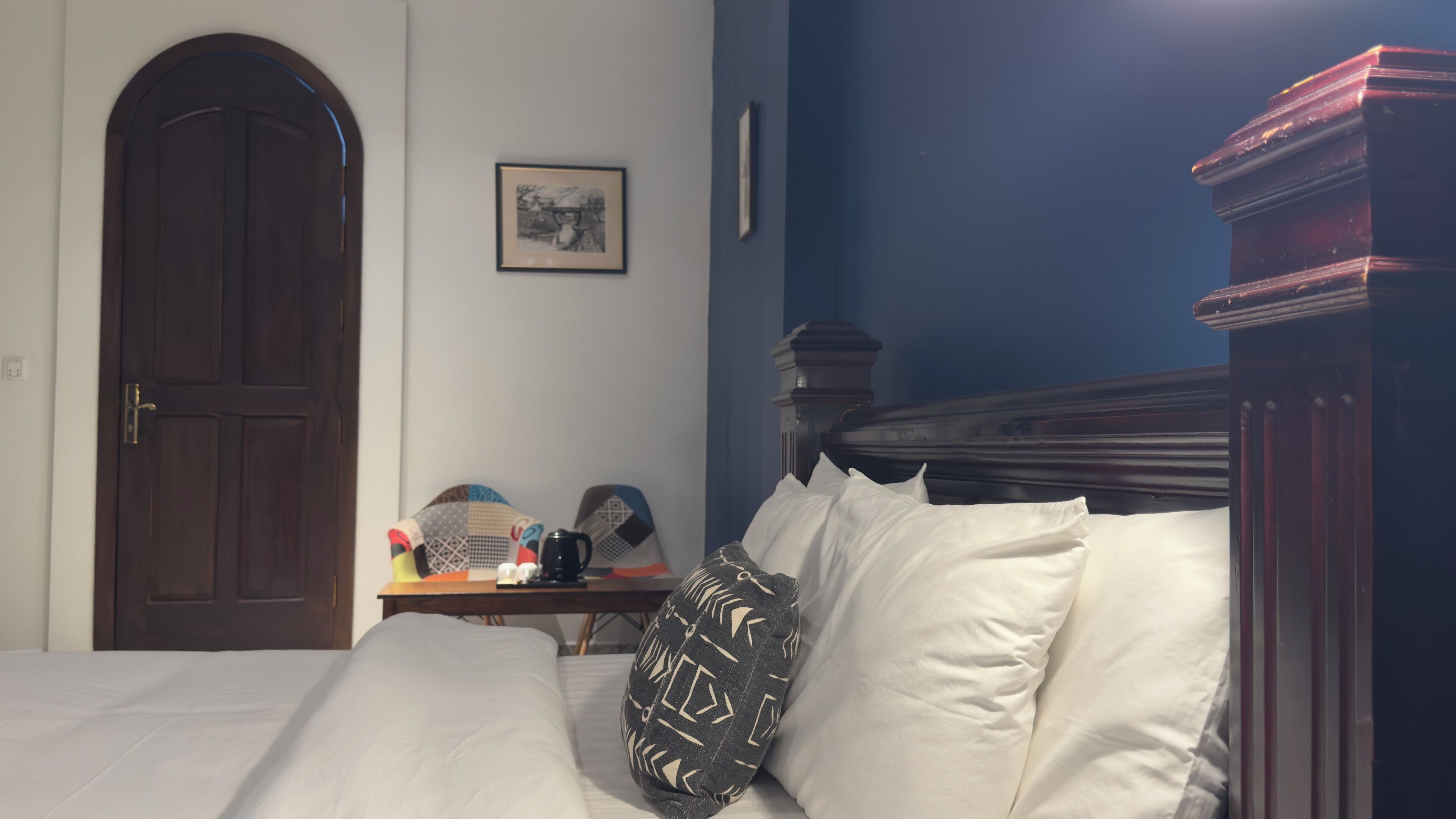 A bedroom with a large bed, a wooden door, a small table with a coffee maker, and a framed picture on the wall.