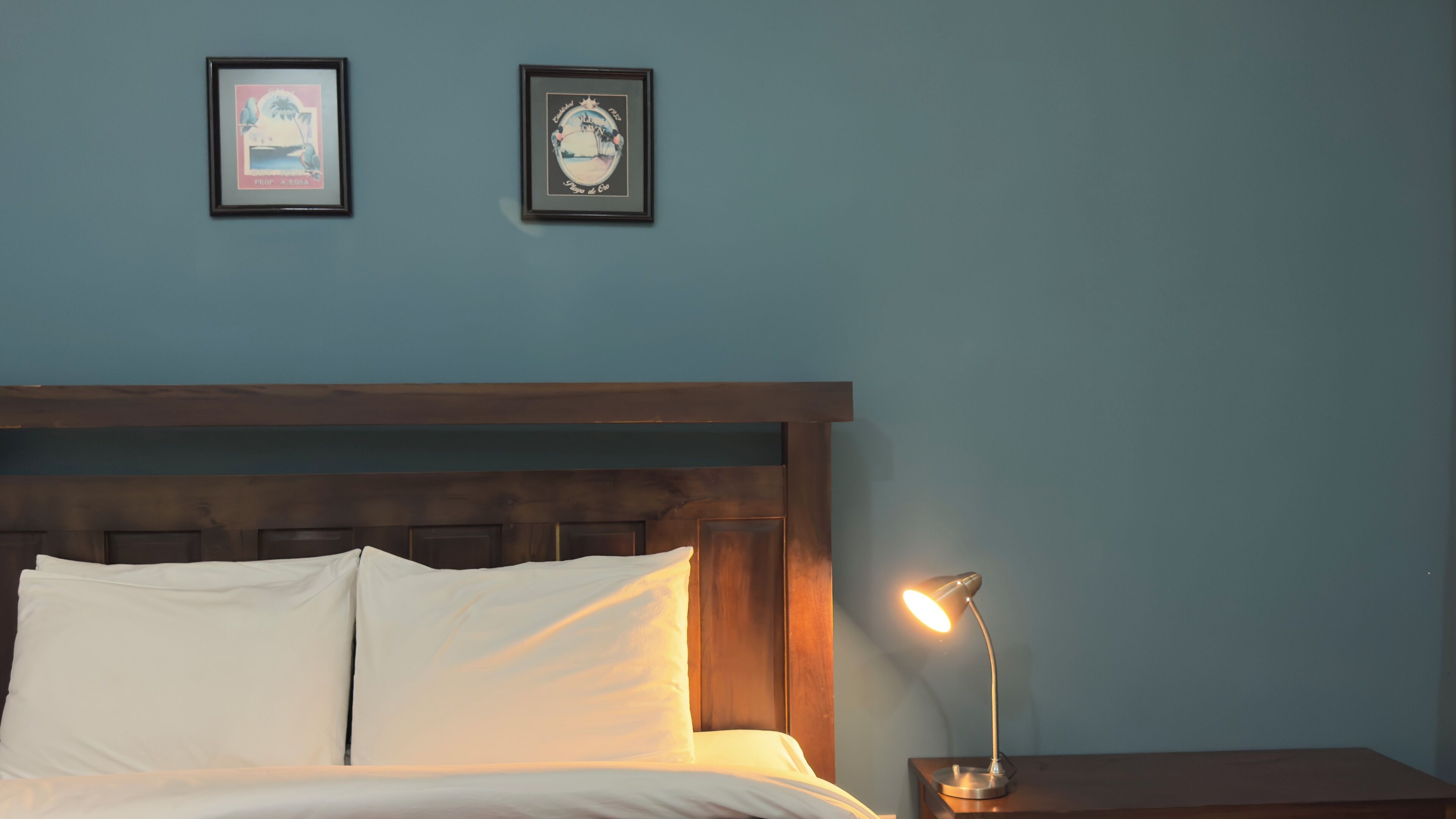 Ein ordentlich bezogenes Bett mit weißen Leinen, ein hölzernes Kopfende und ein Nachttisch mit Lampe. Zwei gerahmte Bilder hängen an der Wand über dem Bett.