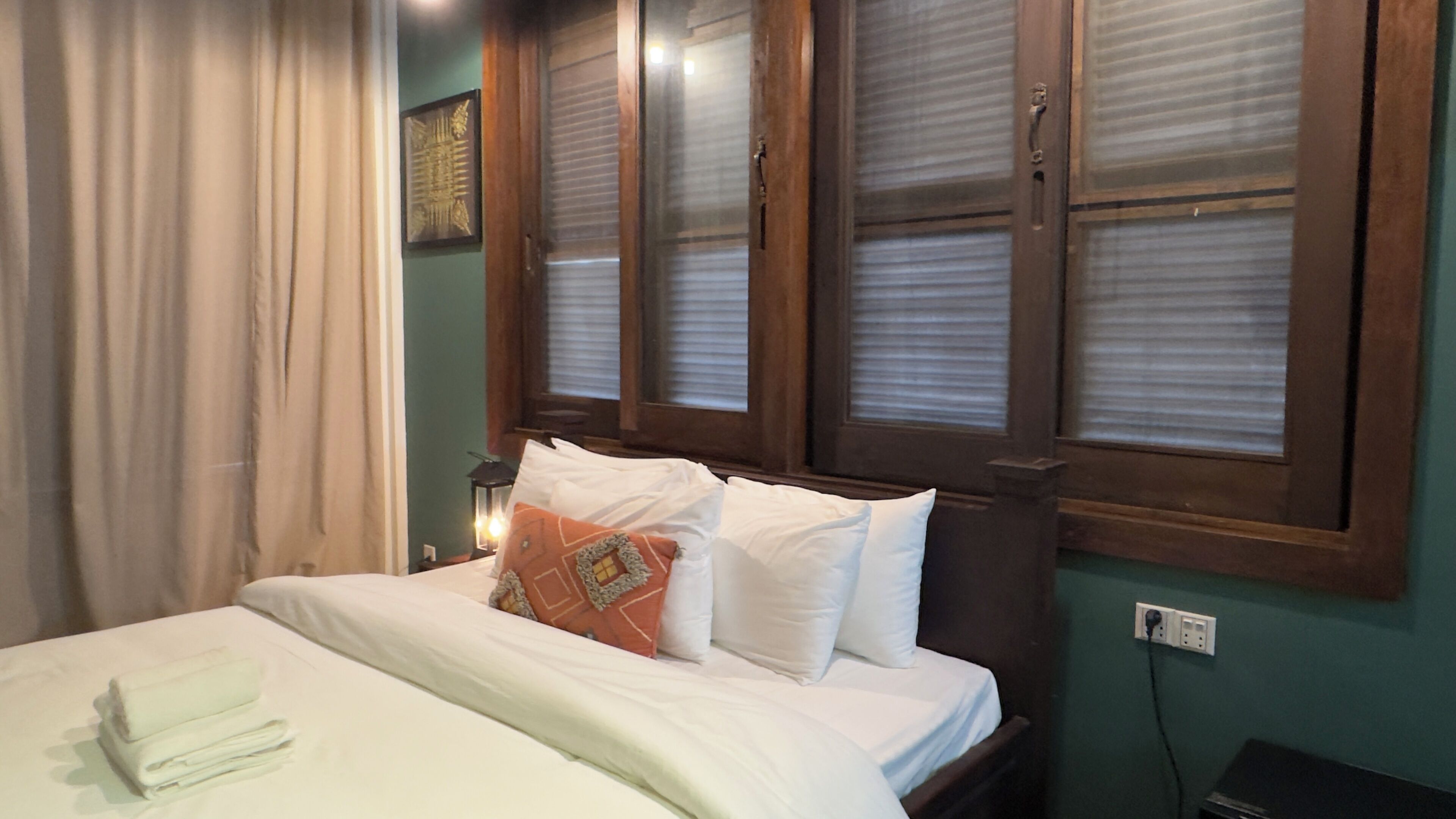 Un lit bien fait, avec du linge de lit blanc et quelques coussins décoratifs, une tête de lit en bois et une fenêtre avec des stores.