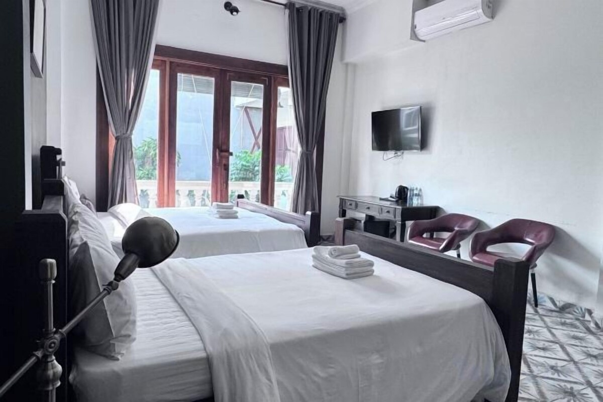 A hotel room with two beds, a desk, a chair, a television, and a balcony with curtains.